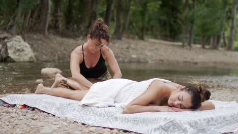 Masseuse-rubbing-feet-of-woman-lying-near-rippling-water-of-pond