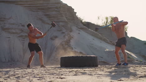 Zwei-Kraftvolle-Männliche-Athleten,-Die-Gemeinsam-Trainieren,-Schlagen-Bei-Sonnenuntergang-In-Der-Wüste-Mit-Einem-Hammer-Auf-Das-Lenkrad.-Ausdauertraining