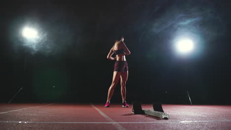 Professional-woman-athlete-on-a-dark-background-gotovtes-to-run-the-sprint-of-Jogging-shoes-in-sneakers-on-the-track-of-the-stadium-on-a-dark-background.-Average-plan