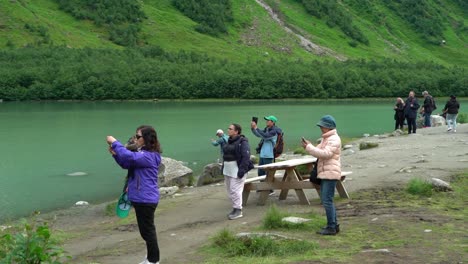 Crowd-of-tourists-standing-on-beach-close-to-green-glacial-lake-and-photographing-Boyabreen-glacier---Slow-motion