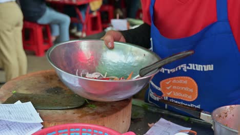 Grabbing-the-condiments-from-the-right-side-of-the-frame,-and-mixing-them-into-a-bowl-to-make-a-vegetable-salad-at-roadside-restaurant-in-the-streets-of-Bangkok,-Thailand