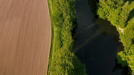 Drone-bird's-eye-view-of-freshly-tilled-field-next-to-wide-river-lined-with-trees