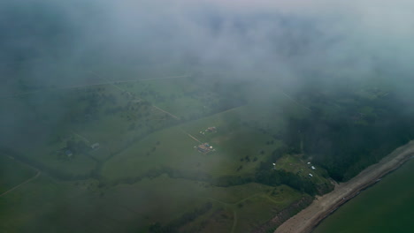 Aerial-drone-shot-over-green-farm-fields-through-rain-clouds-along-rural-countryside-along-the-seaside