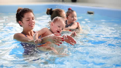 Happy-middle-aged-mother-swimming-with-cute-adorable-baby-in-swimming-pool.-Smiling-mom-and-little-child,-newborn-girl-having-fun-together.-Active-family-spending-leisure-and-time-in-spa-hotel.