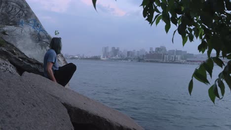 A-Man-in-a-Blue-Shirt-Sitting-on-Some-Rocks-Across-the-River-from-Downtown-Montreal-on-a-Cloudy,-Foggy-Evening