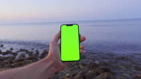 Golden-Hour-POV:-Man's-Hand-Holding-iPhone-14-with-Green-screen-by-Calm-Stoney-Beach,-Chroma-Key