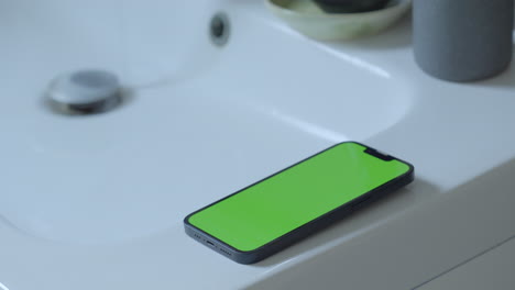 Uso-Creativo:-IPhone-14-Con-Pantalla-Verde-En-El-Lavabo-Del-Baño,-Agua-Que-Gotea-Del-Grifo,-Iluminación-Ambiental,-Croma