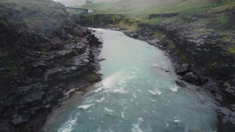 Drone-shot-of-a-raging-Icelandic-river-and-green-fields-above-the-black-rocks-and-rapids