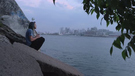 A-Man-in-a-Blue-Shirt-Sitting-on-Some-Rocks-Across-the-River-from-Downtown-Montreal-on-a-Cloudy,-Foggy-Evening