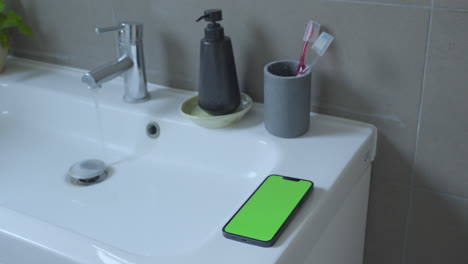 Kreativer-Einsatz:-IPhone-14-Mit-Grünem-Bildschirm-Auf-Dem-Waschbecken-Im-Badezimmer,-Tropfendes-Wasser-Aus-Dem-Wasserhahn,-Umgebungsbeleuchtung,-Chroma-Key
