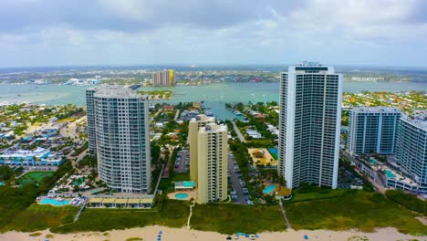 Aerial-drone-shot-slowly-rising-above-the-beach-on-Singer-Island-looking-towards-tall-residential-buildings-in-West-Palm-Beach,-Florida-on-a-blue-sky-day
