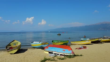 Colorful-boats-on-lake-shore,-people-sunbathing-near-calm-water-in-a-hot-summer-day-in-Pogradec,-Albania