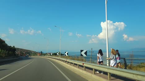 Conduciendo-Por-La-Costa-Del-Lago-Ohrid-En-Una-Carretera-Panorámica,-Nubes-Blancas-Reflejándose-En-La-Superficie-Del-Agua-Tranquila