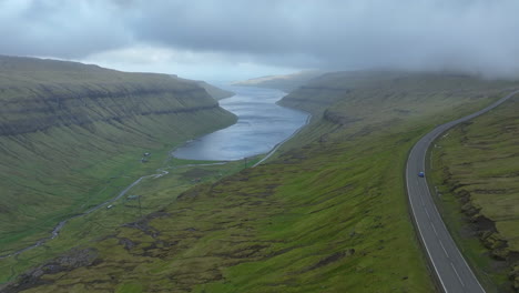 Färöer-Inseln:-Luftaufnahme-Mit-Verfolgung-Eines-Auf-Der-Straße-Fahrenden-Autos,-Wo-Sie-Die-Schönheit-Der-Grünen-Landschaft-Und-Der-Nahegelegenen-Fjorde-Genießen-Können