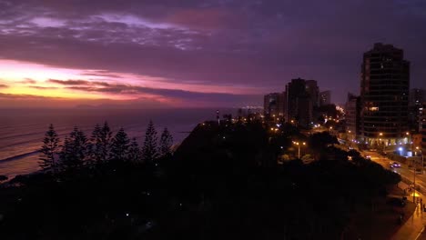 Aerial-shot-shows-a-beautiful-sunset-in-a-lighthouse-park-with-pine-trees-close-to-a-boulevard-where-are-people-walking-next-to-a-street-with-cars-and-buildings-in-a-latin-american-coastal-city