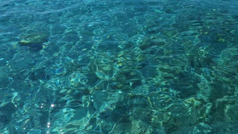 Turquoise-sea-water-background-reflecting-sunlight-through-clean-crystal-water-to-seabed-with-pebbles-and-rocks-in-Ionian-seaside