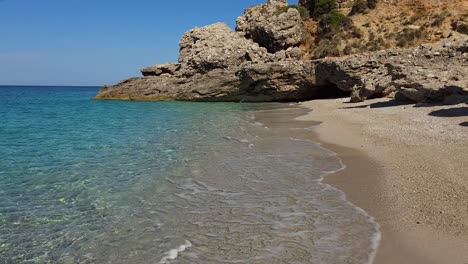 Turquoise-sea-water-washing-untouched-sand-beach-sheltered-by-big-cliffs-on-beautiful-bay-of-Ionian-coastline-in-Albania,-perfect-vacation-spot