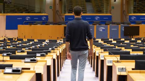 Male-Walking-Inside-Empty-Plenary-Hall-At-The-European-Parliament-And-Sitting-Down-In-Chair