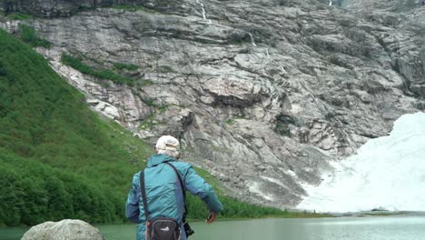 Elderly-tourist-standing-beside-glacial-lake-and-Boyabreen-Norwegian-glacier---Slow-motion-tilt-up-from-person-to-glacier-in-background-mountains