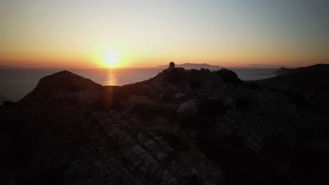 Aerial-orbit-and-reveal-past-rocky-outcropping-at-golden-hour-dusk-as-sun-sets-below-horizon,-syros-greece