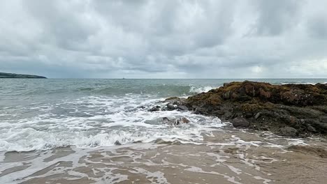 Meereswellen-In-Zeitlupe,-Die-An-Einem-Bewölkten-Morgen-über-Die-Goldenen-Sandstrandfelsen-Der-Insel-Anglesey-Strömen