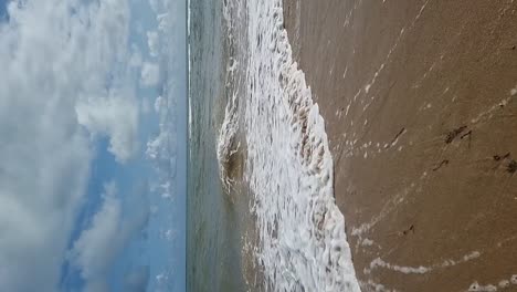 VERTICAL-Tropical-ocean-waves-slow-motion-flowing-across-golden-sandy-beach-under-blue-cloudy-sky