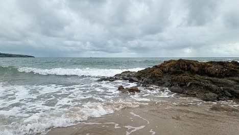 Slow-motion-ocean-waves-washing-across-Anglesey-island-splashing-over-rocks-on-golden-sandy-beach,-overcast-morning