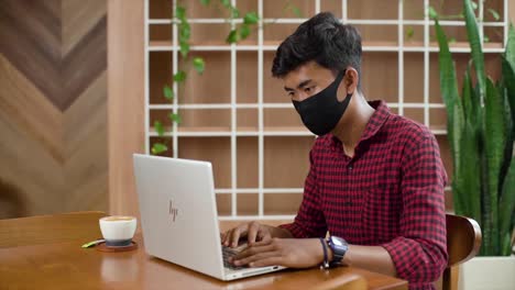 Masked-man-using-laptop-in-cafe-while-sitting-at-wooden-table-and-drink-coffee