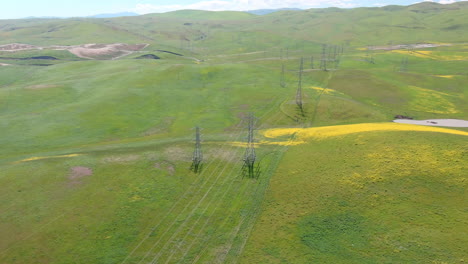 Power-lines-cut-through-idyllic-rolling-green-hills-dotted-with-yellow-wildflowers,-aerial
