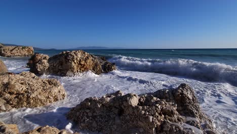 Waves-crash-over-rocks,-splashing-across-cliffs,-violent-breaking-wave-foaming-over-rocky-shoreline-in-Ionian-sea,-Albania