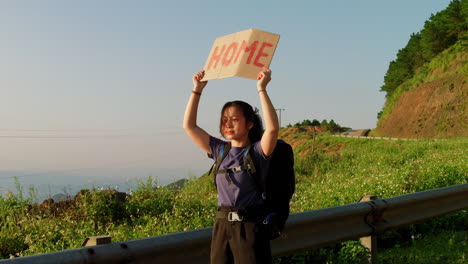 young-solo-female-traveller-hitchhiking-on-road-side-with-a-sign-on-cardboard-saying-home