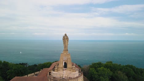 Christus-statue-overlooks-city-of-San-Sebastian-Spain,-drone-dolly-fly-over-to-open-ocean-and-green-coastal-cliff