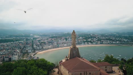 San-Sebastian-Christusstatue-Mit-Blick-Auf-Den-Atemberaubenden-Stadthafen-In-Spanien,-Filmischer-Luftwagen-Mit-Hochfliegenden-Vögeln-Am-Bewölkten-Himmel