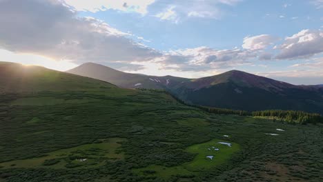 Drone-flyover-calm-alpine-pool-puddles-surrounded-by-vibrant-green-grass-and-small-trees,-sunset-golden-hour-flare-and-colorado-mountain-ridges