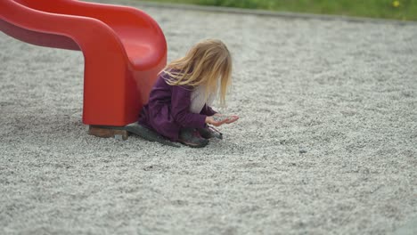 Ein-Blondes-Mädchen-Sitzt-Neben-Der-Rutsche-Auf-Dem-Modernen-Spielplatz-Im-Freien-Und-Spielt-Mit-Steinen