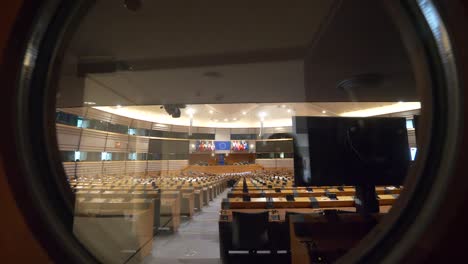 POV-Pull-Back-Shot-From-Round-Glass-Window-Showing-Empty-Plenary-Hall-At-The-European-Parliament-Looking-Towards-Main-Stage-Located-In-Brussels,-Belgium