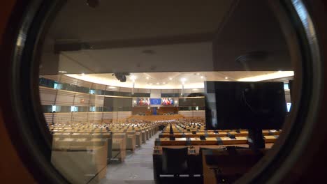 POV-Toma-Hacia-Atrás-Desde-Una-Ventana-Redonda-De-Vidrio-Que-Muestra-La-Sala-De-Plenos-Vacía-Del-Parlamento-Europeo-Mirando-Hacia-El-Escenario-Principal-Ubicado-En-Bruselas,-Bélgica