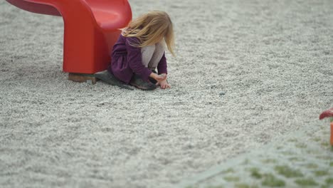 Una-Niña-Rubia-Se-Sienta-Cerca-Del-Tobogán-En-El-Patio-De-Juegos-Al-Aire-Libre-Y-Juega-Con-Piedras