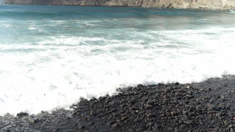 Waves-crashing-against-pebble-black-volcanic-beach-in-Tenerife,-white-foam
