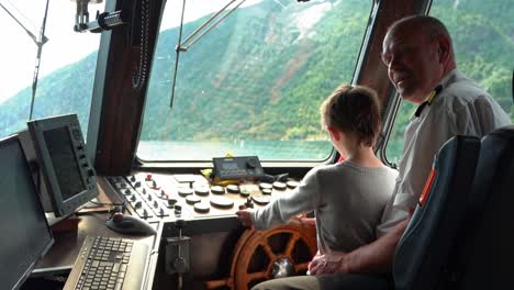 Boy-sitting-on-captains-lap-onboard-ship-and-is-allowed-to-try-steering-with-the-helmsman-wheel