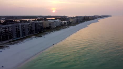 Desting-Florida-Morning-Drone-aerial-view-zooming-in-over-Paradise-Beach-sunrise,-while-the-sun-in-distance-behind-the-building-of-hotel-resort-on-the-beach