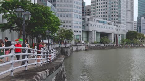 Pedestrians-walk-along-Boat-Quay-with-modern-Skyscrapers-lining-the-Singapore-River,-Singapore