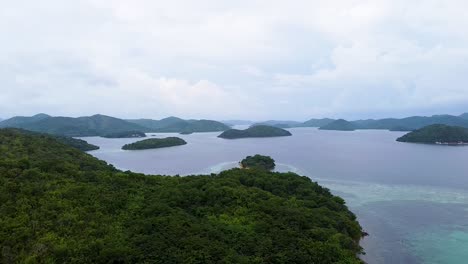 Aerial-drone-lowering-toward-tropical-island-covered-in-trees-and-rainforest-with-scattered-islands-in-remote-Coron-Bay,-Palawan,-Philippines