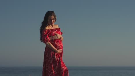 Mittlere-Aufnahme-Einer-Schwangeren-Frau-Mit-Einem-Roten-Kleid,-Die-Ihren-Bauch-Am-Meer-Hält