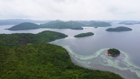 Aerial-view-of-remote-tropical-island-covered-in-trees-and-surrounded-by-coral-reef-ecosystem-in-Coron-Bay,-Palawan,-Philippines,-Southeast-Asia
