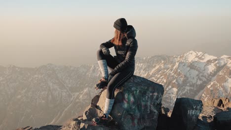Young-caucasian-woman-sitting-on-a-rock-on-top-of-Mount-Toubkal,-with-mountain-range-in-the-background,-Morocco