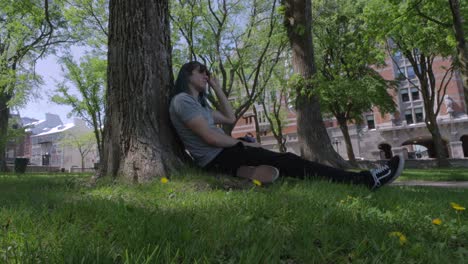 A-Guy-With-Long-Hair-Sitting-and-Relaxing-Under-a-Tree-in-a-Park-in-Quebec-City,-Canada,-on-a-Bright,-Summer-Afternoon