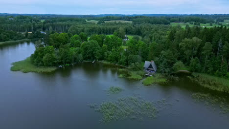 Aerial-Shot-with-Push-in-Low-Angle-Shot-over-a-Lake-with-Views-of-a-Countryside-House-Nestled-Amongst-a-Green-Forest-in-Latvia
