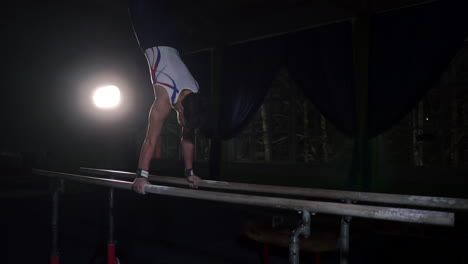 Gimnasta-Profesional-En-Una-Habitación-Oscura-Con-Ropa-Deportiva-Se-Para-Sobre-Cangrejos-De-Río-En-Barras-Paralelas-Y-Realiza-Giros-En-Cámara-Lenta.-Duro-Entrenamiento-En-Preparación-Para-Los-Juegos-Olímpicos.