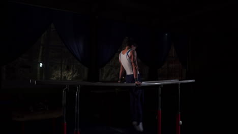 Male-gymnast-acrobat-performs-handstand-on-parallel-bars-in-a-dark-room-in-slow-motion-sharing-a-somersault-and-landing-on-the-floor.-Training-before-the-Olympic-games.-A-professional-athlete
